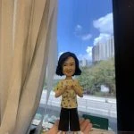 客製化Q版女士公仔——比心手勢休閒造型 手工人物公仔細節 （圖片展示了一尊客製化的Q版女性人物黏土公仔，主角留著及肩的黑色微捲短髮（Bob頭），氣質優雅，面帶溫柔微笑。她身穿一件黃色底配白色與綠色碎花圖案的短袖上衣，下身搭配黑色寬腳褲（Culottes），腳踩黑色尖頭高跟鞋，造型時尚且休閒。公仔最獨特的動作是雙手在胸前比出「愛心」手勢，展現出親切、充滿愛意與關懷的形象。公仔站立在一個簡約的白色圓形底座上。） 環境背景與光影 （公仔由一隻手托起展示，背景位於室內窗邊。左側有垂下的米白色窗簾，窗外是大片蔚藍的天空、白雲以及遠處的城市樓景與綠樹，還有下方的高架道路。光線充足且自然，明亮的日光讓公仔的膚色與衣著色彩顯得格外清晰自然。下方工作檯上可見橙色美工刀、畫筆等製作工具，顯示這是在工作室內完成後的實拍記錄。） 軟陶公仔、真人公仔訂製、Q版人像、女士禮物、比心手勢、休閒造型、手工禮物、黃色碎花衫、寬腳褲穿搭、生日禮物 表達愛意、溫馨紀念、感謝心意、母親節禮物、週年紀念、獨一無二、辦公室擺設、幸福時刻