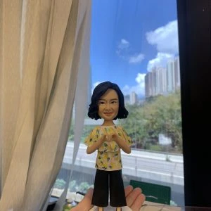 客製化Q版女士公仔——比心手勢休閒造型 手工人物公仔細節 （圖片展示了一尊客製化的Q版女性人物黏土公仔，主角留著及肩的黑色微捲短髮（Bob頭），氣質優雅，面帶溫柔微笑。她身穿一件黃色底配白色與綠色碎花圖案的短袖上衣，下身搭配黑色寬腳褲（Culottes），腳踩黑色尖頭高跟鞋，造型時尚且休閒。公仔最獨特的動作是雙手在胸前比出「愛心」手勢，展現出親切、充滿愛意與關懷的形象。公仔站立在一個簡約的白色圓形底座上。） 環境背景與光影 （公仔由一隻手托起展示，背景位於室內窗邊。左側有垂下的米白色窗簾，窗外是大片蔚藍的天空、白雲以及遠處的城市樓景與綠樹，還有下方的高架道路。光線充足且自然，明亮的日光讓公仔的膚色與衣著色彩顯得格外清晰自然。下方工作檯上可見橙色美工刀、畫筆等製作工具，顯示這是在工作室內完成後的實拍記錄。） 軟陶公仔、真人公仔訂製、Q版人像、女士禮物、比心手勢、休閒造型、手工禮物、黃色碎花衫、寬腳褲穿搭、生日禮物 表達愛意、溫馨紀念、感謝心意、母親節禮物、週年紀念、獨一無二、辦公室擺設、幸福時刻