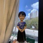 客製化Q版女士公仔——短髮藍衫優雅造型 手工人物公仔細節 （圖片展示了一尊客製化的Q版女性人物黏土公仔，主角留著俐落的黑色短髮，面容慈祥，嘴角帶著淺淺的微笑。她身穿一件藍色短袖上衣，衣服上有獨特的白色流線型花紋及袖口處的黃色格紋裝飾，下身搭配黑色長裙，腳踩黑色鞋子，整體造型端莊大方。公仔的雙手自然交疊於腹前，展現出嫻淑、穩重且彬彬有禮的姿態。公仔站立在一個簡約的白色圓形底座上。） 環境背景與光影 （公仔由一隻手托起展示，背景位於室內窗邊。左側有垂下的米白色窗簾，窗外是大片蔚藍的天空、白雲以及遠處的城市樓景與綠樹，還有下方的高架道路。光線充足且自然，讓公仔的面部表情與衣著細節清晰可見。下方工作檯上隱約可見橙色美工刀等製作工具，顯示這是作品完成後的實拍記錄。） 軟陶公仔、真人公仔訂製、Q版人像、長輩禮物、短髮造型、藍色上衣、黑色長裙、端莊站姿、手工禮物、榮休禮物 表達敬意、溫馨紀念、感謝師恩、母親節禮物、退休紀念、獨一無二、優雅氣質、珍貴回憶