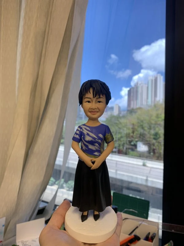 客製化Q版女士公仔——短髮藍衫優雅造型 手工人物公仔細節 （圖片展示了一尊客製化的Q版女性人物黏土公仔，主角留著俐落的黑色短髮，面容慈祥，嘴角帶著淺淺的微笑。她身穿一件藍色短袖上衣，衣服上有獨特的白色流線型花紋及袖口處的黃色格紋裝飾，下身搭配黑色長裙，腳踩黑色鞋子，整體造型端莊大方。公仔的雙手自然交疊於腹前，展現出嫻淑、穩重且彬彬有禮的姿態。公仔站立在一個簡約的白色圓形底座上。） 環境背景與光影 （公仔由一隻手托起展示，背景位於室內窗邊。左側有垂下的米白色窗簾，窗外是大片蔚藍的天空、白雲以及遠處的城市樓景與綠樹，還有下方的高架道路。光線充足且自然，讓公仔的面部表情與衣著細節清晰可見。下方工作檯上隱約可見橙色美工刀等製作工具，顯示這是作品完成後的實拍記錄。） 軟陶公仔、真人公仔訂製、Q版人像、長輩禮物、短髮造型、藍色上衣、黑色長裙、端莊站姿、手工禮物、榮休禮物 表達敬意、溫馨紀念、感謝師恩、母親節禮物、退休紀念、獨一無二、優雅氣質、珍貴回憶
