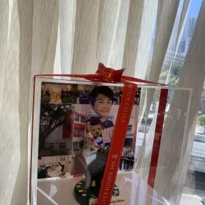 一座透明禮盒包裝的桌面公仔擺設,外繫紅色緞帶與金色字樣。盒內中央是一位短髮微笑的人像公仔,穿深色禮袍式披肩與長裙,雙手捧著泰迪熊與一張迷你證書/卡片;腳邊是綠色圓形草地底座,散落白粉色小花。背景板拼貼多張校園相關照片,包含紅白相間外牆的校舍、校園樹蔭、辦公室場景與團體合照。底部白色標牌印有英文名稱與“Principal”字樣。整體放置在手掌上,背後是室內米色窗簾與窗外城市景致。