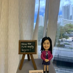 一座放在手掌上的桌面模型：右側是一個穿紫粉色外套、深藍碎花連身裙與黑色鞋子的女性人像公仔，雙手交握微笑站立；長直髮、戴耳環。左側是一個木質畫架小黑板，上面以白色字體寫著「Now the FUN really begins!」。底座為灰色顆粒紋地台，前方白色小牌標示「HTCSS 2024」。背景是窗外的城市高樓與藍天白雲，室內有米色窗簾。