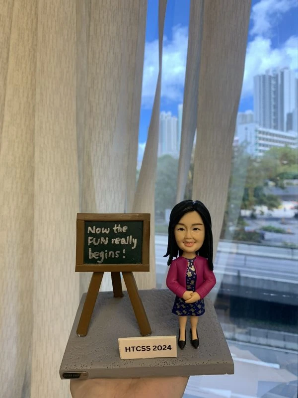 一座放在手掌上的桌面模型：右側是一個穿紫粉色外套、深藍碎花連身裙與黑色鞋子的女性人像公仔，雙手交握微笑站立；長直髮、戴耳環。左側是一個木質畫架小黑板，上面以白色字體寫著「Now the FUN really begins!」。底座為灰色顆粒紋地台，前方白色小牌標示「HTCSS 2024」。背景是窗外的城市高樓與藍天白雲，室內有米色窗簾。