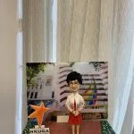 手托一座小型桌面場景模型：中央是一位短髮、戴眼鏡的人像公仔，穿白色外套搭配彩色花卉印花袖口與紅色連身裙，雙手於腹前交握站立。背景立牌印有一所學校建築照片，立面帶有紅色水平線與彩虹色斜紋裝飾，四周樹葉遮蔭。左前方立著一顆橙色星形標誌與「HKUGA PRIMARY SCHOOL」字樣的小牌。底座為紅棕色磚紋地面，兩側綠色帶狀區域點綴白色小花；前方金屬銘牌寫著「HKUGA Primary School Principal Wong Kwai Ling」。