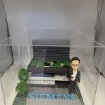 一座企業主題 diorama：右側是身著黑色西裝與白襯衫、戴眼鏡的人像公仔，右手握小獎盃；中央至後景為層次分明的灰黑色建築模型，屋頂佈置綠植區塊與步道；前方地面以灰磚鋪設，放置醒目藍色“SIEMENS”立體字，左前方有樹與花草點綴，整體封裝於透明壓克力盒中。