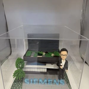 一座企業主題 diorama:右側是身著黑色西裝與白襯衫、戴眼鏡的人像公仔,右手握小獎盃;中央至後景為層次分明的灰黑色建築模型,屋頂佈置綠植區塊與步道;前方地面以灰磚鋪設,放置醒目藍色“SIEMENS”立體字,左前方有樹與花草點綴,整體封裝於透明壓克力盒中。