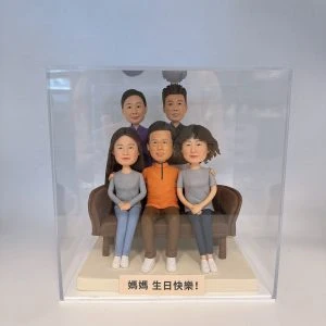 一組家庭主題的人像公仔擺設放在透明展示盒內:前排三人坐在咖啡色沙發上,左側長髮女生與右側短髮女生穿灰色上衣與運動鞋,中間男子穿橙色上衣與眼鏡;後排兩位站在後方,一位穿紫色上衣、一位穿深色上衣。底座前方有淺色小牌寫著「媽媽 生日快樂!」。背景是簡潔的室內白色環境,展示盒邊緣與反光可見。