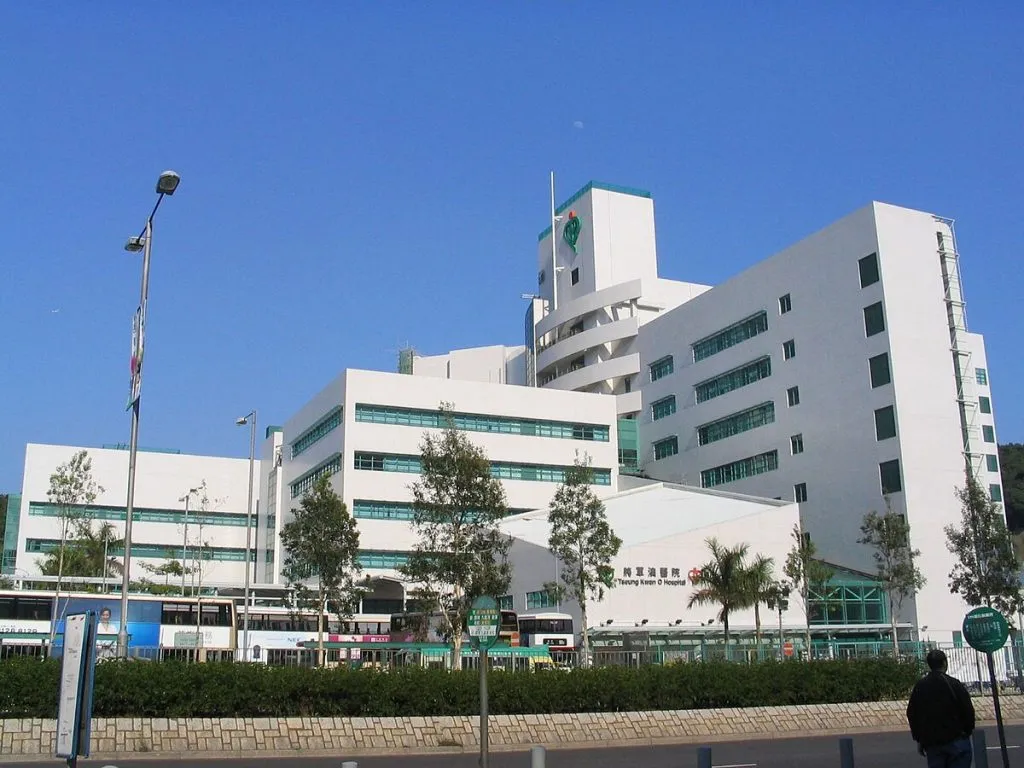 將軍澳醫院的白色建築物在藍天下，前面有巴士站和行人，是紀念將軍澳醫院顧問護師的背景。