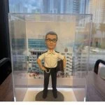 透明展示盒內。公仔角色留著短髮，佩戴眼鏡，身穿整齊的白色制服襯衫與黑色長褲，腰間配有繩帶，左手自然垂落並拿著外套，右手叉腰站立，神情自信莊重。腳下為灰色圓形底座，正面設有淺金色小名牌。背景背板為救護總隊大樓外牆照片，具有鮮明的職業象徵與紀念意義。整體設計將專業形象、退休榮譽與珍重祝福融為一體，莊重中帶溫情，極具收藏價值。