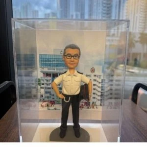 透明展示盒內。公仔角色留著短髮，佩戴眼鏡，身穿整齊的白色制服襯衫與黑色長褲，腰間配有繩帶，左手自然垂落並拿著外套，右手叉腰站立，神情自信莊重。腳下為灰色圓形底座，正面設有淺金色小名牌。背景背板為救護總隊大樓外牆照片，具有鮮明的職業象徵與紀念意義。整體設計將專業形象、退休榮譽與珍重祝福融為一體，莊重中帶溫情，極具收藏價值。