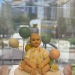 透明展示盒內，一款坐姿的BB人像公仔擺放在白色圓形底座上；娃娃穿著帶有黃色花紋與花領的淺色上衣，雙腳盤坐、雙手自然放在膝上。身後以藍、綠、黃、粉等馬卡龍色氣球作背景，底座側邊點綴雛菊與粉花。前方名牌以中英文字寫著「Gianna 歡迎你」。整體在窗邊自然光下呈現，遠景可見城市高樓與藍天倒影。