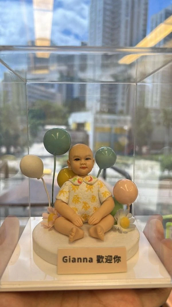 透明展示盒內，一款坐姿的BB人像公仔擺放在白色圓形底座上；娃娃穿著帶有黃色花紋與花領的淺色上衣，雙腳盤坐、雙手自然放在膝上。身後以藍、綠、黃、粉等馬卡龍色氣球作背景，底座側邊點綴雛菊與粉花。前方名牌以中英文字寫著「Gianna 歡迎你」。整體在窗邊自然光下呈現，遠景可見城市高樓與藍天倒影。