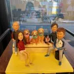 一組全家福人像公仔與狗狗公仔的迷你場景：四位角色圍坐在階梯式座台兩側與前方，中央台階上並排四隻狗狗，包括三隻金色長毛犬配綠色與粉色領巾，以及一隻黑白棕配色的犬隻戴粉紅掛牌。人物服裝各異：左側為紅色上衣與白色短裙、後方站著穿背心的長者；右側為藍白運動外套與運動鞋的年輕人，旁邊還有一位戴眼鏡的角色。整體置於黃色底板與木質桌面上，背後是窗景。