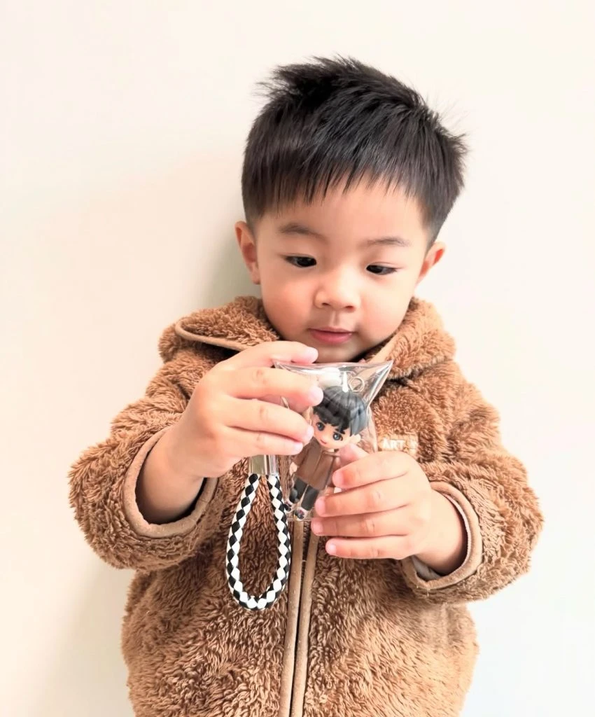 近距離視角，小朋友雙手高舉透明吊袋，袋中可見同款Q版人像公仔與金屬圈扣，毛絨外套質感清晰。