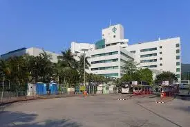 將軍澳醫院外觀，一棟白色建築物在藍天白雲下，前方有行車道和棕櫚樹，象徵著對醫院顧問護師的致敬與榮譽。