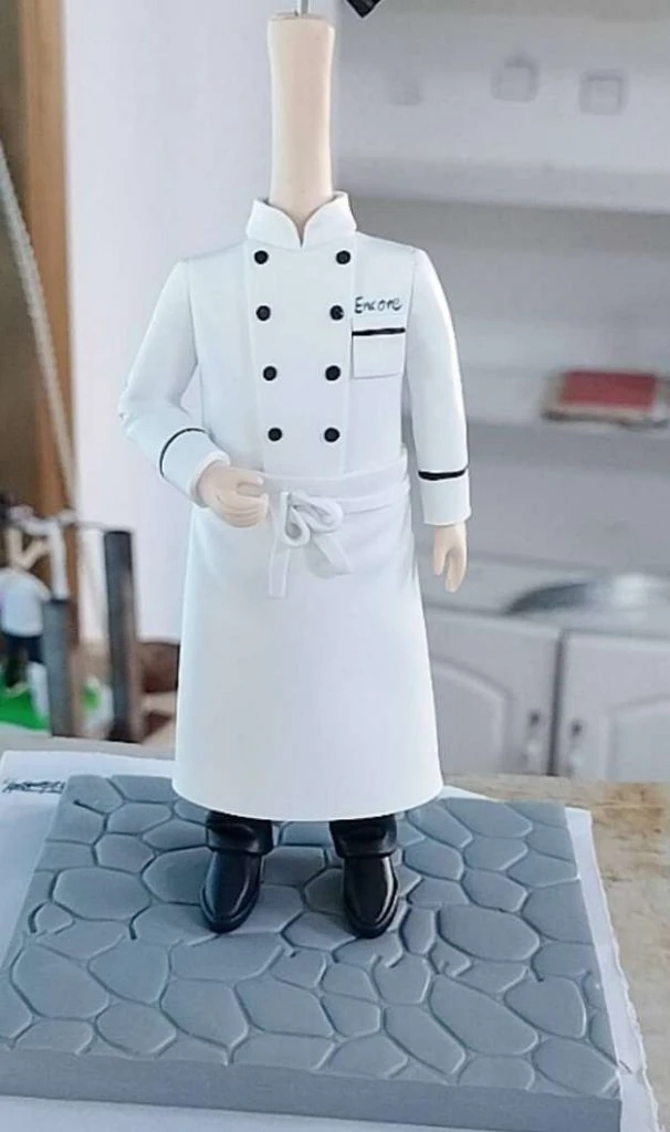 半成品人像公仔：只見身體未裝頭部，穿白色雙排扣廚師服與圍裙，胸前寫有「Encore」名牌，黑色長褲與皮鞋站在灰色石板紋底座上，右手微彎、左手自然下垂。