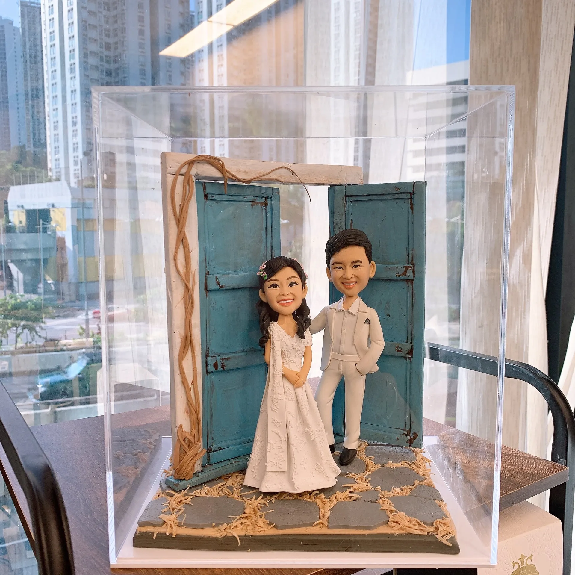 透明展示盒中的婚禮公仔場景：新娘穿白色蕾絲禮服站在做舊的藍色木門前，新郎穿全白西裝一手插袋站在旁邊，腳下是石板路與乾草裝飾，窗外可見城市高樓景色。