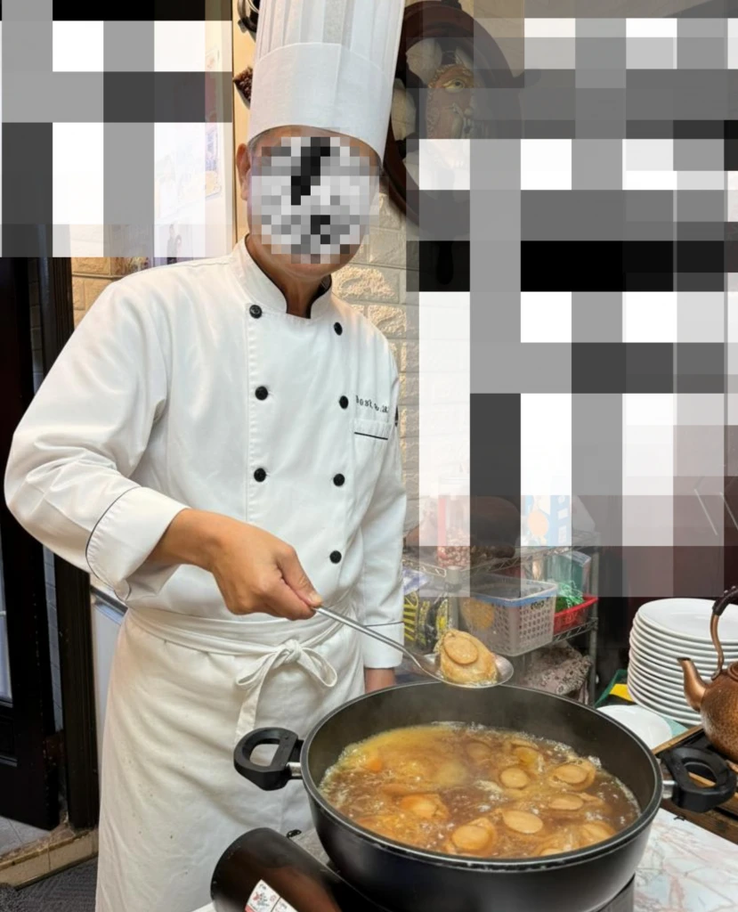廚師身穿白色雙排扣廚師服與高帽，腰間綁帶，右手持湯勺從大鍋中撈起鮑魚；背景為餐廳內部與餐具。臉部以馬賽克遮蔽。