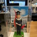 男畢業生主題人像公仔穿深藍學袍戴學士帽，手持文憑站在草地底座與校園背景前，透明展示盒內附金色名牌