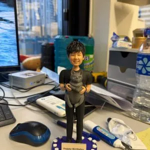 男子公仔抱著灰色貓咪站在紫色愛心底座，上緣環繞白色小玫瑰並配有「Happy Birthday」金色銘牌，生日寵物主題客製