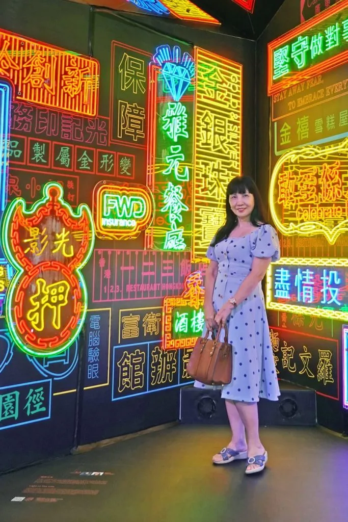 一位女士穿著藍色波點裙，手挽咖啡色手袋，站立於色彩繽紛的香港霓虹招牌前，展示城市記憶的浪漫童話。