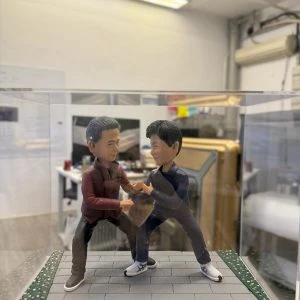 透明展示盒內的雙人公仔：兩名男子在灰色磚道地台上對練站樁交手，左側穿酒紅外套黑褲，右側著深灰運動套裝與白色武術鞋，地台邊緣有草皮與碎花點綴。