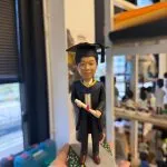 男畢業生人像公仔穿黑色學袍與方帽，手持紅綁帶文憑，站在灰色步道與綠地小花底座上，典禮感十足