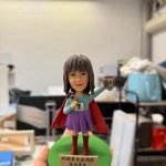 英雄披風女店長公仔高舉咖啡杯與字牌，綠色底座附祝賀銘牌，開幕與業績慶祝客製禮