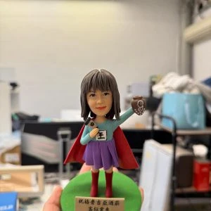 英雄披風女店長公仔高舉咖啡杯與字牌，綠色底座附祝賀銘牌，開幕與業績慶祝客製禮