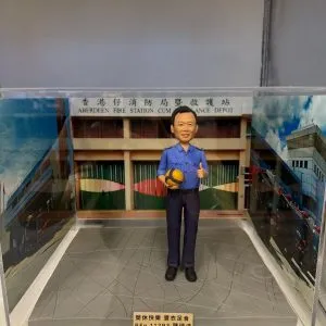 透明展示盒內的人像公仔：身穿藍色制服的男子手抱黃藍排球，站在灰色地台上；背板為「香港仔消防局暨救護站」建築外觀，前方金色銘牌刻有祝福語與編號。