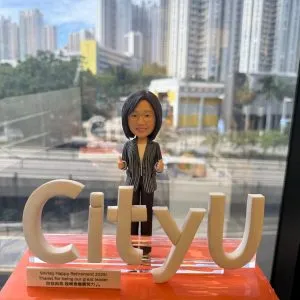 女性人像公仔穿條紋西裝外套站於橙色亮面底座上，前方以立體「CityU」字樣做造型，側前方貼有英中雙語退休祝福銘牌