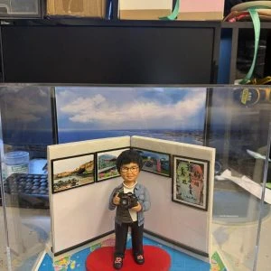 透明展示盒中的攝影主題公仔：短髮戴眼鏡的人物手持相機站在紅色愛心地墊上，背後三折屏風掛有風景照與書法字畫；地台鋪世界地圖，前方銘牌寫「Tracy 海闊天空」。