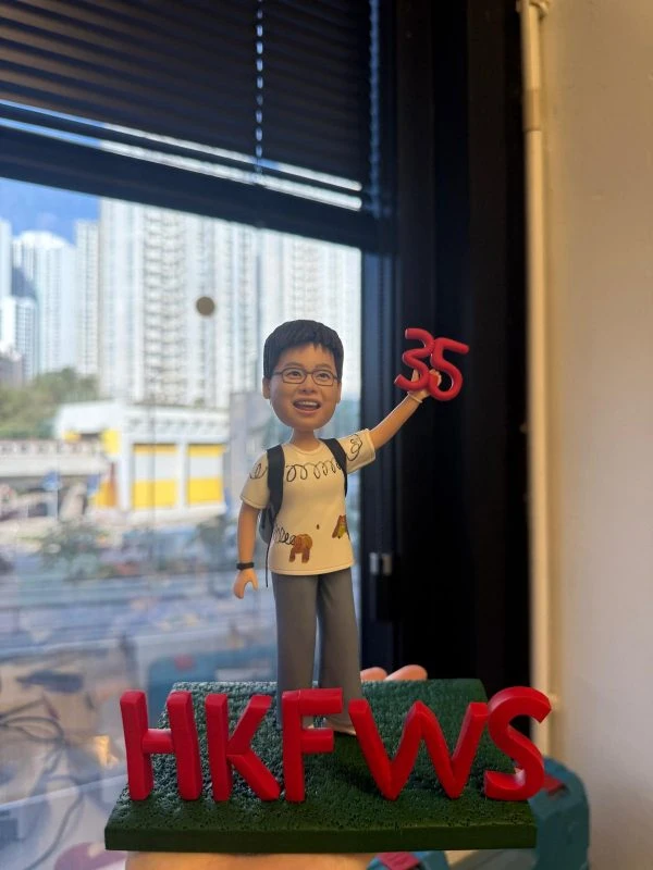 戴眼鏡的人像公仔背著背包、穿白色塗鴉T與灰色長褲，右手高舉紅色「35」，站在綠色草地底座，前方立體紅字「HKFWS」