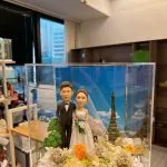 一對婚禮人像公仔站在花藝佈置的草地上，身穿黑色禮服與白色婚紗手捧花束，後方是藍天白雲與迷你巴黎鐵塔元素，整體置於透明展示盒內並隱藏暖黃小燈