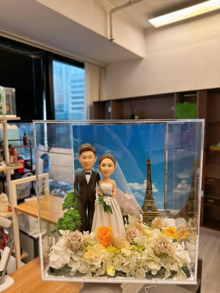 一對婚禮人像公仔站在花藝佈置的草地上，身穿黑色禮服與白色婚紗手捧花束，後方是藍天白雲與迷你巴黎鐵塔元素，整體置於透明展示盒內並隱藏暖黃小燈
