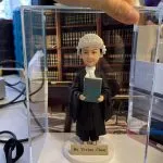 女大律師人像公仔戴傳統白色法庭假髮與黑色法袍，手持綠色法律手冊，置於透明展示盒內，前方金色銘牌刻 Ms Vivian Chan，背景為書牆律所場景