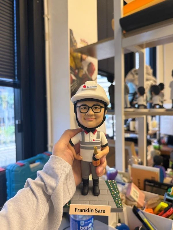 工程師主題人像公仔戴白色安全帽與對講耳機，穿公司POLO制服站在工地磚紋底座，手持香檳並配有姓名銘牌 Franklin Sare