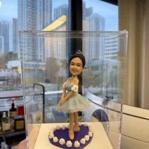 透明展示盒內的芭蕾公仔：女孩戴小皇冠，穿淡藍色薄紗舞裙與芭蕾舞鞋，雙手置前微屈膝擺出優雅姿勢；腳下為紫色愛心底座，邊緣圍繞白色玫瑰裝飾。