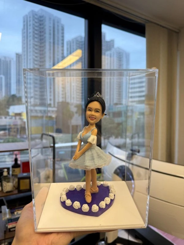 透明展示盒內的芭蕾公仔：女孩戴小皇冠，穿淡藍色薄紗舞裙與芭蕾舞鞋，雙手置前微屈膝擺出優雅姿勢；腳下為紫色愛心底座，邊緣圍繞白色玫瑰裝飾。