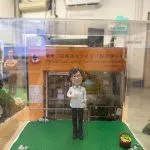 人像公仔穿白色制服站在綠色草地底座上，背後是「東華三院鄒蓮女士長者日間護理中心」門面背板，前方放有刻字銘牌，旁邊點綴小石與菜籃