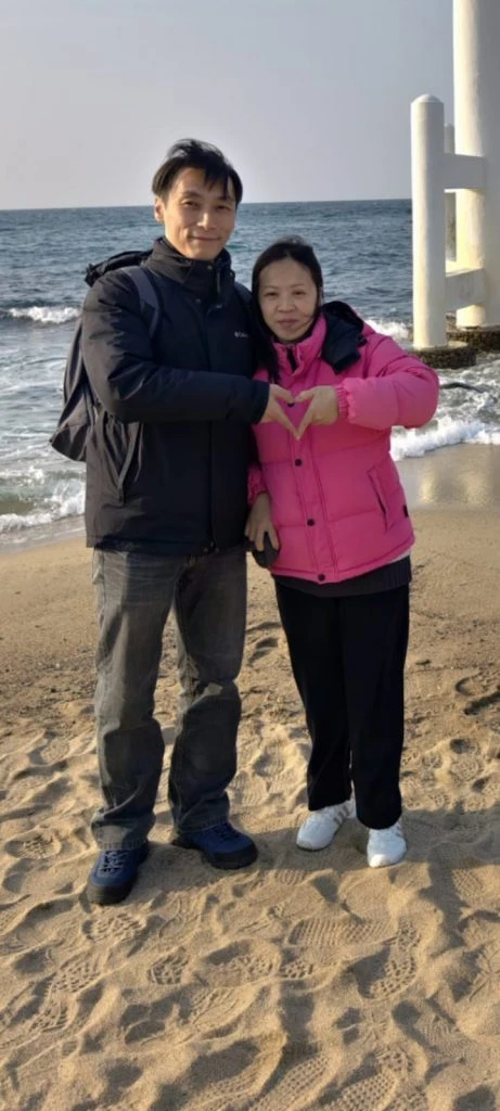 一對情侶在沙灘上合照，女士穿粉紅色羽絨外套並用雙手比出心形，男士穿深藍色外套，象徵客製化公仔能將瞬間快照化為永恆場景。