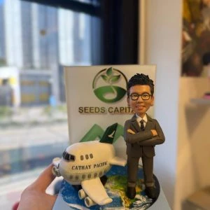 西裝人像公仔雙手交叉站在地球圖案底座旁，旁邊有印上 CATHAY PACIFIC 的小客機模型，後方立體背板寫有 SEEDS CAPITAL 與綠色標誌