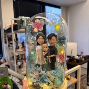 三人家庭人像公仔置於木座玻璃罩內,周圍以藍綠與粉色系花材環繞並點綴暖黃燈,呈現溫馨合照氛圍