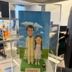 親子雙人公仔站在綠色草地底座上，後方是藍天白雲背板與白色小椅，前方銘牌刻字「April happy birthday」，旁邊擺有小盆栽與石頭點綴