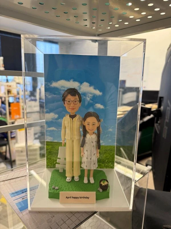 親子雙人公仔站在綠色草地底座上，後方是藍天白雲背板與白色小椅，前方銘牌刻字「April happy birthday」，旁邊擺有小盆栽與石頭點綴