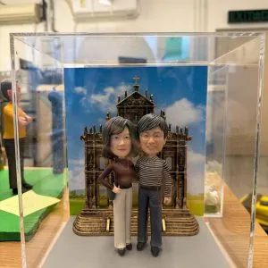 兩位人像公仔親密站立於灰色底座上,背後是澳門大三巴立體背板與藍天白雲圖,前方銘牌寫上金婚紀念字樣,整體置於透明展示盒內