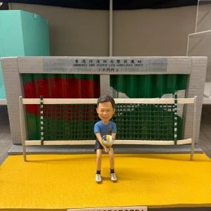 公仔坐在黃綠相間的排球門前，手抱排球，身穿藍色上衣與黑色短褲；背板為「香港仔消防局暨救護站」車房捲門，前方有致敬銘牌。