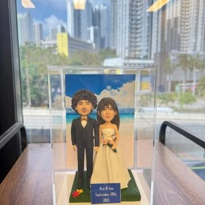 透明展示盒中的婚禮公仔：新郎著黑色西裝與蝴蝶結，新娘穿無肩白紗手捧粉橘花束，腳下綠草地台，背板為藍天白雲與海岸；前方小牌寫著「Keri & Tom September 20th, 2025」。