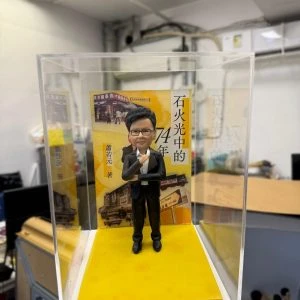 身穿西裝的手工軟陶單人公仔,置於透明展示盒內,背景為黃色書封場景,展現精緻雕塑與立體收藏效果。