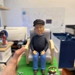 長者人像公仔戴報童帽、穿深色外套與藍褲，坐在木扶手椅上，置於綠色草地底座，旁邊有裝著蔬菜的小籃與石頭點綴，神情和藹